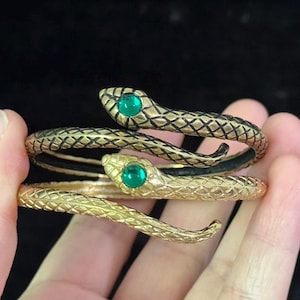 Könnte beinhalten: Goldfarbene Schlangen-Armreifen mit smaragdgrünen Augen. Die schlangenförmigen Armbänder haben ein strukturiertes Schuppenmuster. Der Schmuck wird vor einem dunklen Hintergrund präsentiert.