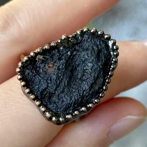 May include: A dark, irregularly shaped ring with a textured, black stone. The ring's setting is a dark metal with a beaded border. The ring is shown on a finger.