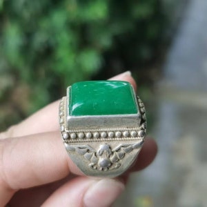 May include: A silver ring with a large, square, bright green stone. The ring has a detailed design around the stone and on the band, featuring a decorative emblem. The ring is held in a hand.