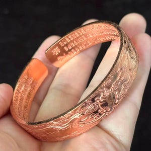 May include: Copper-colored open cuff bracelet with engraved Chinese characters and a detailed dragon design. The bracelet is held in a hand, showcasing the intricate patterns and warm metallic tones. A unique piece of jewelry.
