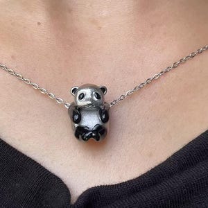 May include: A silver-colored panda pendant necklace. The panda has black accents on its ears, eyes, and limbs. The necklace chain is also silver. The panda is the focal point of the necklace.