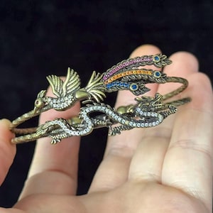 May include: A brass-toned cuff bracelet featuring a detailed phoenix and dragon design. The phoenix has colorful gemstone accents on its tail feathers, while the dragon body is encrusted with clear crystals. The band has a textured, rope-like finish.