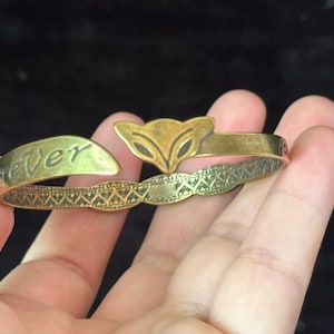 Puede incluir: Un brazalete de latón con un diseño de cabeza de zorro. El brazalete tiene un diseño abierto con la palabra "ever" grabada en un extremo. La banda tiene un patrón decorativo. El brazalete se sostiene en una mano.