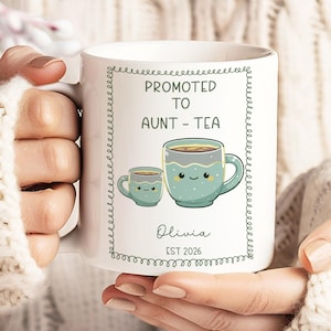 May include: White ceramic mug featuring the text "PROMOTED TO AUNT - TEA" and a charming illustration of two teacups. The mug also includes the name "Olivia" and the year "EST 2026". The mug is held by a person wearing a white sweater.