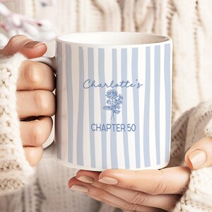 以下が含まれることがあります： 水色の縦縞模様の白いセラミックマグカップ。筆記体で「Charlotte's CHAPTER 50」と書かれています。白いニットのセーターを着た人がマグカップを持っています。