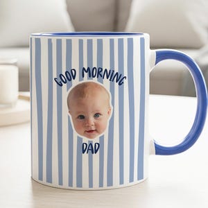 May include: A ceramic mug featuring a light blue and white vertical striped pattern. The front displays a photographic portrait of a baby centered between the dark blue text that reads Good Morning Dad in a simple, rounded sans-serif font.