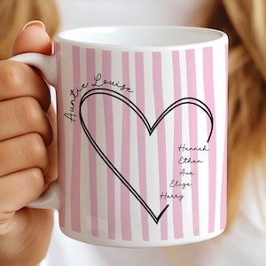 May include: White ceramic mug with pink and white vertical stripes. A black heart design is centered on the mug, with the names "Auntie Louise" and four other names written in black script. The mug is held by a person.