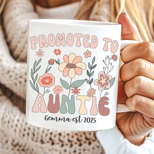 May include: White ceramic mug featuring the text "PROMOTED TO AUNTIE" in a retro font, with floral illustrations. The mug also includes the text "Gemma est 2025".