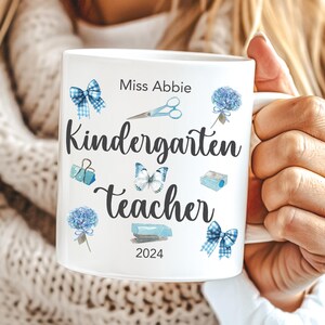 Op de afbeelding: Witte keramische mok met de tekst "Kindergarten Teacher" en de naam "Miss Abbie". De mok is versierd met blauwe aquarel illustraties van strikken, scharen, vlinders en andere schoolgerelateerde elementen. Het jaar "2024" staat ook op de mok.