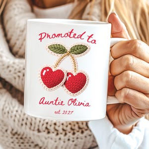 May include: A white ceramic mug featuring a graphic of two red heart-shaped cherries with green leaves. The text reads Promoted to Auntie Olivia est 2027 in red script. The design mimics a knitted texture with a light-coloured border around the cherries.