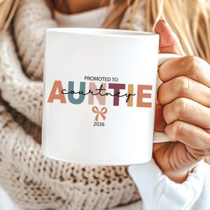 May include: White ceramic mug with the text "Promoted to Auntie Courtney 2026" in colourful letters. A small bow graphic is below the text. The mug is held by a person wearing a white sweater and a cream-coloured scarf.