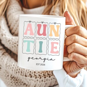 May include: White ceramic mug featuring the word "AUNTIE" in a retro, colourful design. The name "georgia" is written in cursive below, with "EST 2026" underneath. The mug is held by a person wearing a white sweater and a cream-coloured scarf.