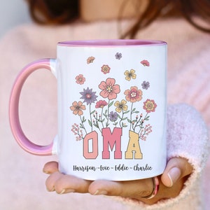 Puede incluir: Taza de cerámica blanca con asa e interior rosas. La taza presenta la palabra "OMA" en letras de bloque rosas y amarillas, rodeada de un diseño floral en varios colores. Debajo de la palabra "OMA" están los nombres "Harrison - Evie - Eddie - Charlie".