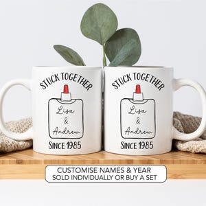 Op de afbeelding: Twee witte keramische mokken met de woorden "STUCK TOGETHER" boven een lijmflesontwerp. De namen "Lisa & Andrew" en het jaar "SINCE 1985" staan eronder gedrukt. De mokken staan op een houten oppervlak.