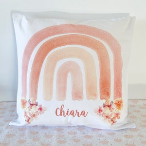 May include: A white pillow with a watercolor rainbow design in shades of peach, orange, and pink. The rainbow is accented with small white flowers. The name "Chiara" is printed in cursive below the rainbow.