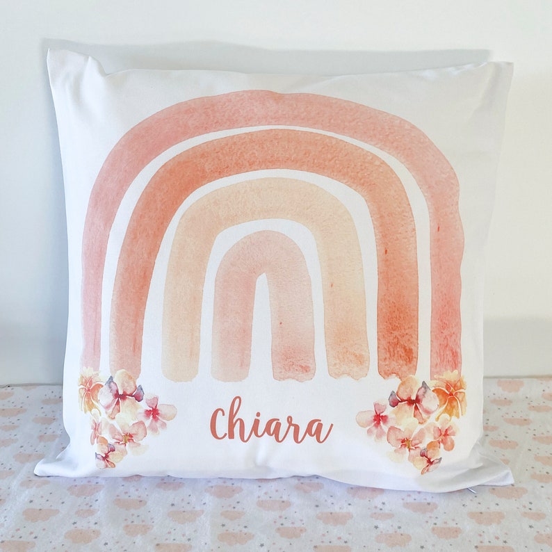 May include: A white pillow with a watercolor rainbow design in shades of peach, orange, and pink. The rainbow is accented with small white flowers. The name "Chiara" is printed in cursive below the rainbow.