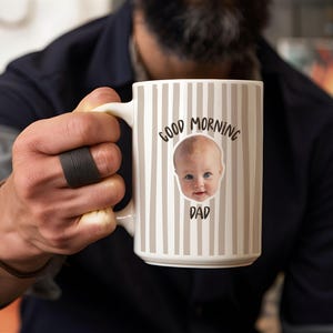 May include: A white ceramic mug featuring a vertical beige striped pattern. The front of the mug displays a photographic image of a baby's face centered between the black text Good Morning above and the black text DAD below the baby's face.