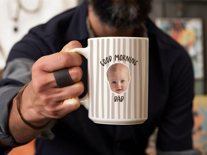 May include: A white ceramic mug featuring a vertical beige striped pattern. The front of the mug displays a photographic image of a baby's face centered between the black text Good Morning above and the black text DAD below the baby's face.
