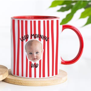 May include: A white ceramic mug with a red vertical striped pattern, a red interior, and a red handle. The front features a photographic print of a baby's face centered between the black text "GOOD MORNING" above and "DAD" below the image.