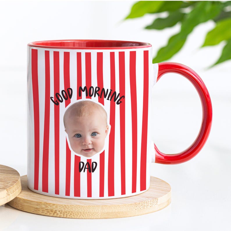 May include: A white ceramic mug with a red vertical striped pattern, a red interior, and a red handle. The front features a photographic print of a baby's face centered between the black text "GOOD MORNING" above and "DAD" below the image.
