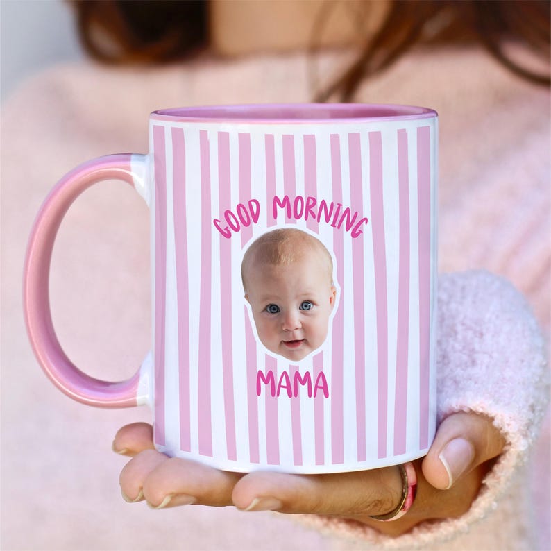May include: A ceramic mug with a pink and white vertical striped pattern. The mug features a pink handle and interior. Centered on the front is a photographic portrait of a baby's face with the pink text GOOD MORNING MAMA printed above and below.