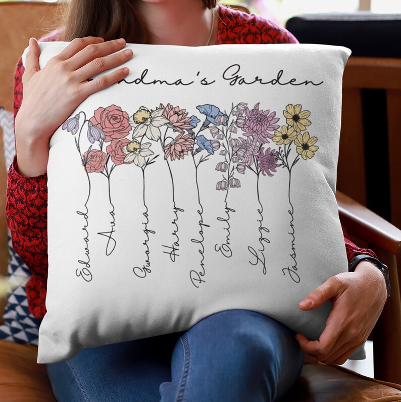 May include: White cushion with the text "Grandma's Garden" printed in black cursive script. The cushion features a floral design with seven different flowers, each with a name written underneath: Edward, Ava, Georgia, Harry, Penelope, Emily, Lizzie, and Jasmine.