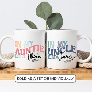 May include: Two white ceramic mugs with curved handles. One mug reads "IN MY AUNTIE ERA Olivia est2004" in colourful text. The other mug reads "IN MY UNCLE ERA James est2004". A sprig of greenery is behind the mugs.