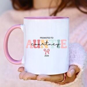 May include: White ceramic mug with a pink interior and handle. The mug features the text "PROMOTED TO Auntie Courtney 2026" in colourful letters and a bow graphic. A perfect gift for a new aunt.
