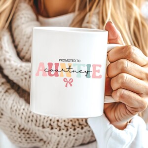 May include: White ceramic mug with the text "PROMOTED TO AUNTIE Courtney" in colourful letters and a small bow. The mug is held by a person.