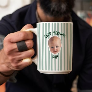 May include: A white ceramic mug featuring a pattern of vertical sage green stripes. The center of the mug displays a photographic image of a baby's face surrounded by the dark green text that reads Good Morning Dad in a curved arrangement.