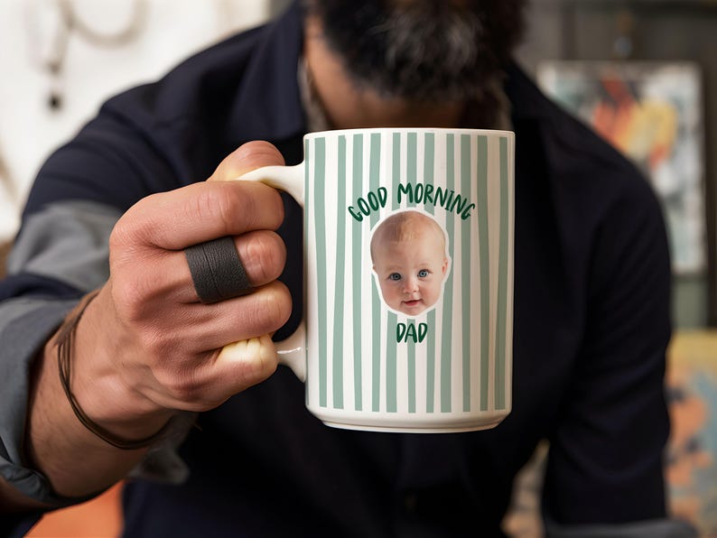 May include: A white ceramic mug featuring a pattern of vertical sage green stripes. The center of the mug displays a photographic image of a baby's face surrounded by the dark green text that reads Good Morning Dad in a curved arrangement.