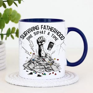 May include: White ceramic coffee mug with a blue rim. The mug features a black and white illustration of a fist breaking through a pile of papers with the text "Surviving Fatherhood One Sip at a Time".