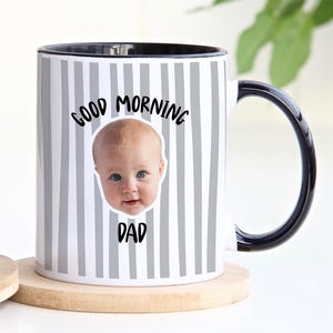 May include: A white ceramic mug with a black handle and interior, featuring a vertical gray striped pattern. The mug displays a photographic print of a baby's face centered between the black text that reads GOOD MORNING DAD in a rounded font.