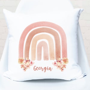 May include: White pillow with a watercolor rainbow in shades of pink, orange, and brown. The rainbow has flowers at the bottom and the name "Georgia" is written below the flowers.