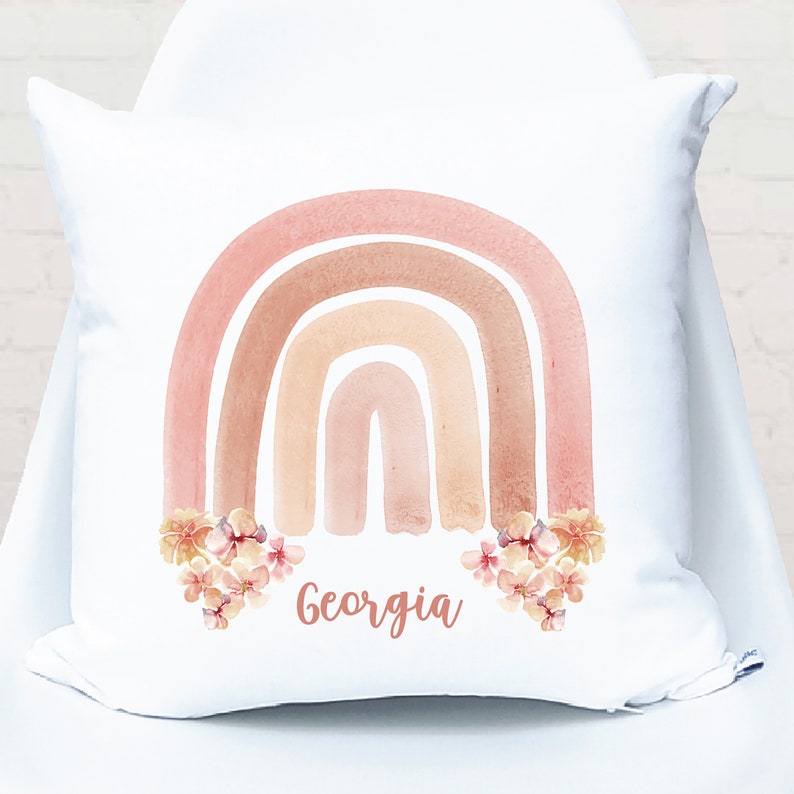 May include: White pillow with a watercolor rainbow in shades of pink, orange, and brown. The rainbow has flowers at the bottom and the name "Georgia" is written below the flowers.