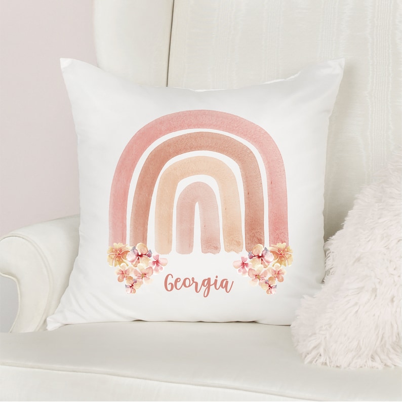 May include: A white pillow with a watercolor rainbow design in shades of pink and brown. The rainbow is decorated with small pink and white flowers. The name "Georgia" is written in black below the rainbow.