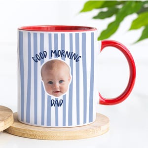 May include: A white ceramic mug with a red interior and handle, featuring a light blue vertical striped pattern. The mug displays a photographic print of a baby's face centered between the dark blue text that reads Good Morning Dad.