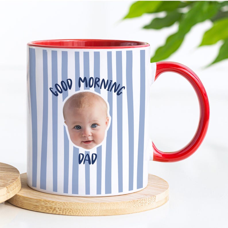May include: A white ceramic mug with a red interior and handle, featuring a light blue vertical striped pattern. The mug displays a photographic print of a baby's face centered between the dark blue text that reads Good Morning Dad.