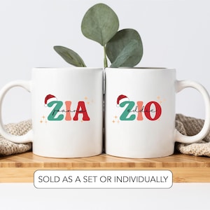 May include: Two white ceramic mugs, each featuring a name in red and teal with a Santa hat design. The names are "Joanna" and "Eddie". The mugs sit on a wooden surface, with greenery behind them. The text at the bottom reads "SOLD AS A SET OR INDIVIDUALLY."