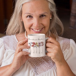 Pu&ograve; includere: Una tazza in ceramica bianca con la scritta "Grandma's Garden" e illustrazioni floreali. La tazza ha nomi sotto i fiori. La tazza &egrave; tenuta da una persona che indossa un top bianco.
