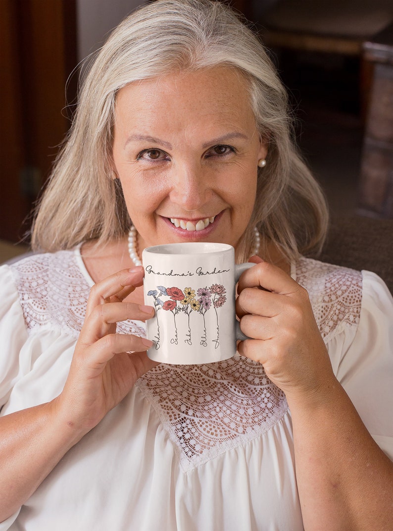 Pu&ograve; includere: Una tazza in ceramica bianca con la scritta "Grandma's Garden" e illustrazioni floreali. La tazza ha nomi sotto i fiori. La tazza &egrave; tenuta da una persona che indossa un top bianco.