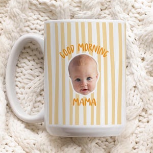 May include: A white ceramic mug featuring a vertical yellow striped pattern. The center of the mug displays a photographic portrait of a baby's face, with the orange text GOOD MORNING arched above the face and the word MAMA printed below it.
