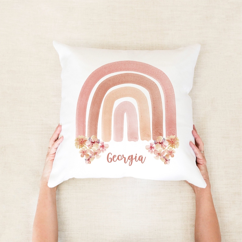 May include: White pillow with a watercolor rainbow design in shades of pink and brown. The rainbow is decorated with pink flowers and the name "Georgia" is written in a cursive font below the rainbow.