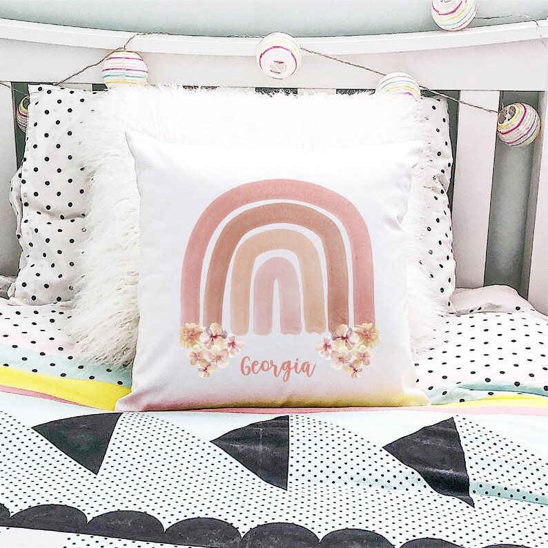May include: A white pillow with a pink, orange, and brown watercolor rainbow design. The rainbow is accented with small white flowers. The name "Georgia" is written in cursive below the rainbow.