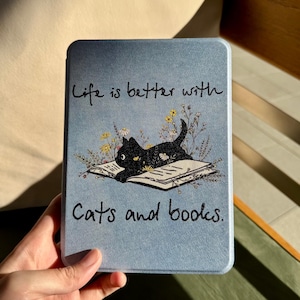 May include: A light blue tablet case featuring a black cat lying on an open book with floral accents. The text "Life is better with Cats and books" is written in black script.