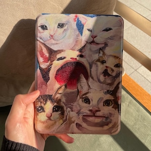 Può includere: Una custodia rettangolare per e-reader con un collage colorato di vari gatti protagonisti di meme su internet. L'illustrazione mostra diversi volti felini con tratti espressivi ed esagerati in uno stile pittorico su tutta la superficie.