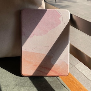Könnte beinhalten: Eine rechteckige Tablethülle mit einem Aquarelldesign in Rosa- und Pfirsichtönen. Ein feines schwarzes Linienkunstdetail ist auf der Hülle zu sehen. Die Hülle liegt auf einer grünen Oberfläche, mit einem beigen Kissen im Hintergrund.