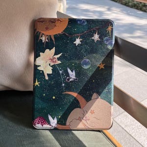 Può includere: Una custodia decorativa con un design celeste, con una luna crescente, stelle e un gatto. La custodia ha uno sfondo verde scuro con un'illustrazione stravagante di un sole, stelle e una figura fatata. La custodia è progettata per proteggere un dispositivo elettronico.
