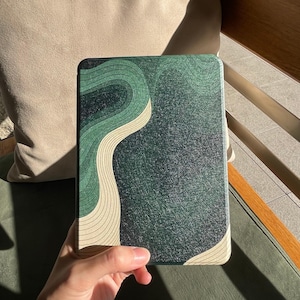 Puede incluir: Una funda rectangular para lector electrónico con una superficie texturizada en verde oscuro y negro, que presenta un patrón fluido de estilo topográfico con líneas de color crema que se curvan por el lado izquierdo y el borde inferior.