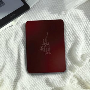 Può includere: Una custodia per e-reader bordeaux con un design a fiamma bianca. La custodia rettangolare con angoli arrotondati è appoggiata su una coperta bianca e strutturata. Un lettore Kindle è parzialmente visibile sullo sfondo.